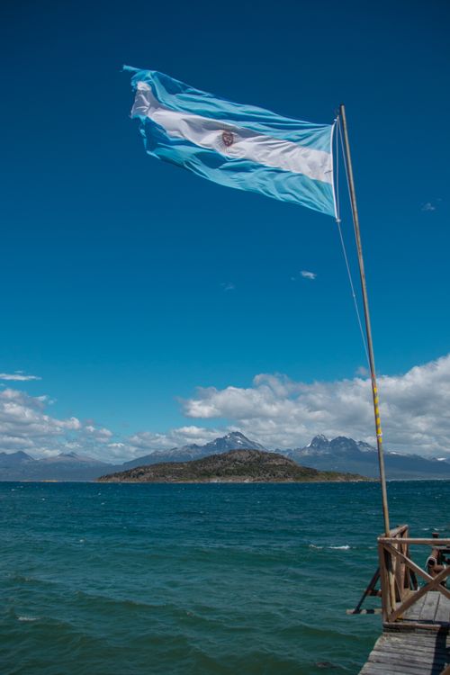 Free A vibrant image of the Argentine flag flying above the blue waters of Ushuaia with mountain views. Stock Photo