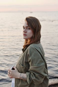 Portrait of a young woman in an olive jacket standing by the ocean during sunset.