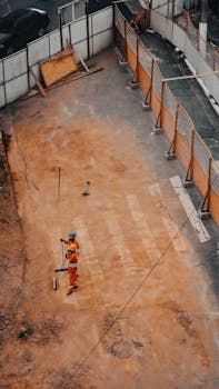 Construction worker on site, viewed from above, wearing safety gear. Daylight setting with visible equipment.