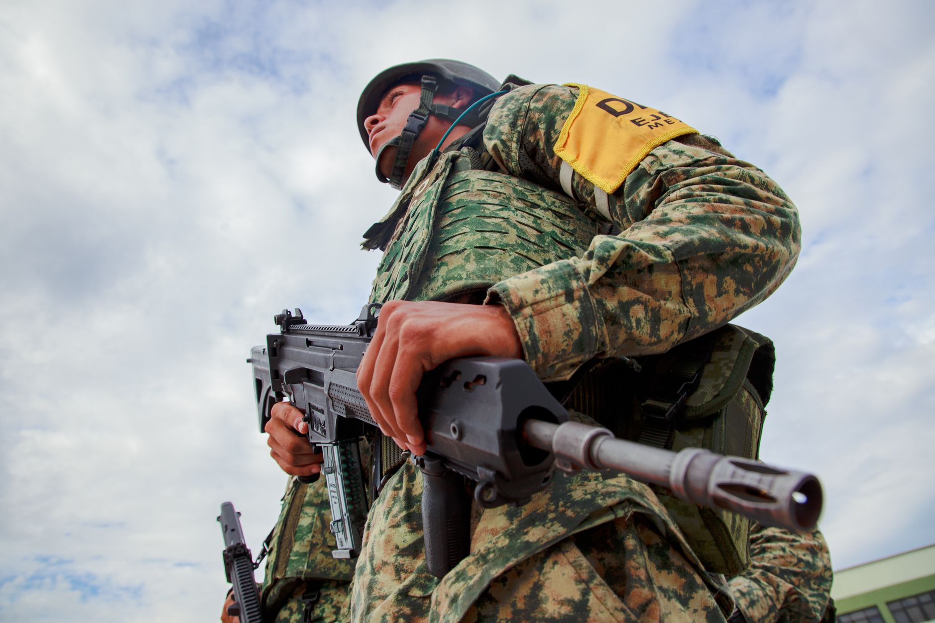 Close-up of a soldier in camouflage holding a rifle under a blue sky, representing military readiness in Mexico.