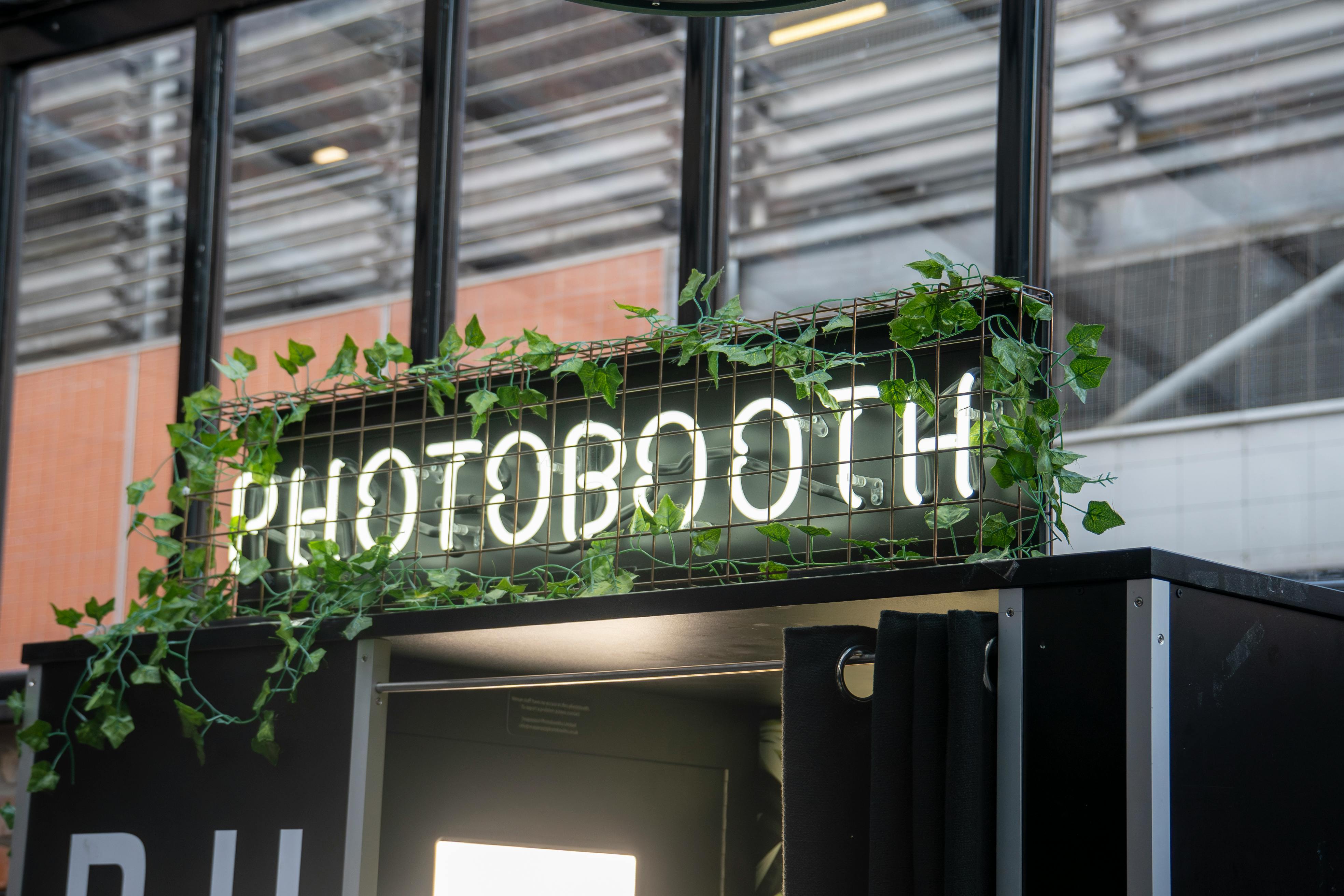 Photobooth au design contemporain avec enseigne lumineuse et végétation, parfait pour une personnalisation de marque lors d'événements.