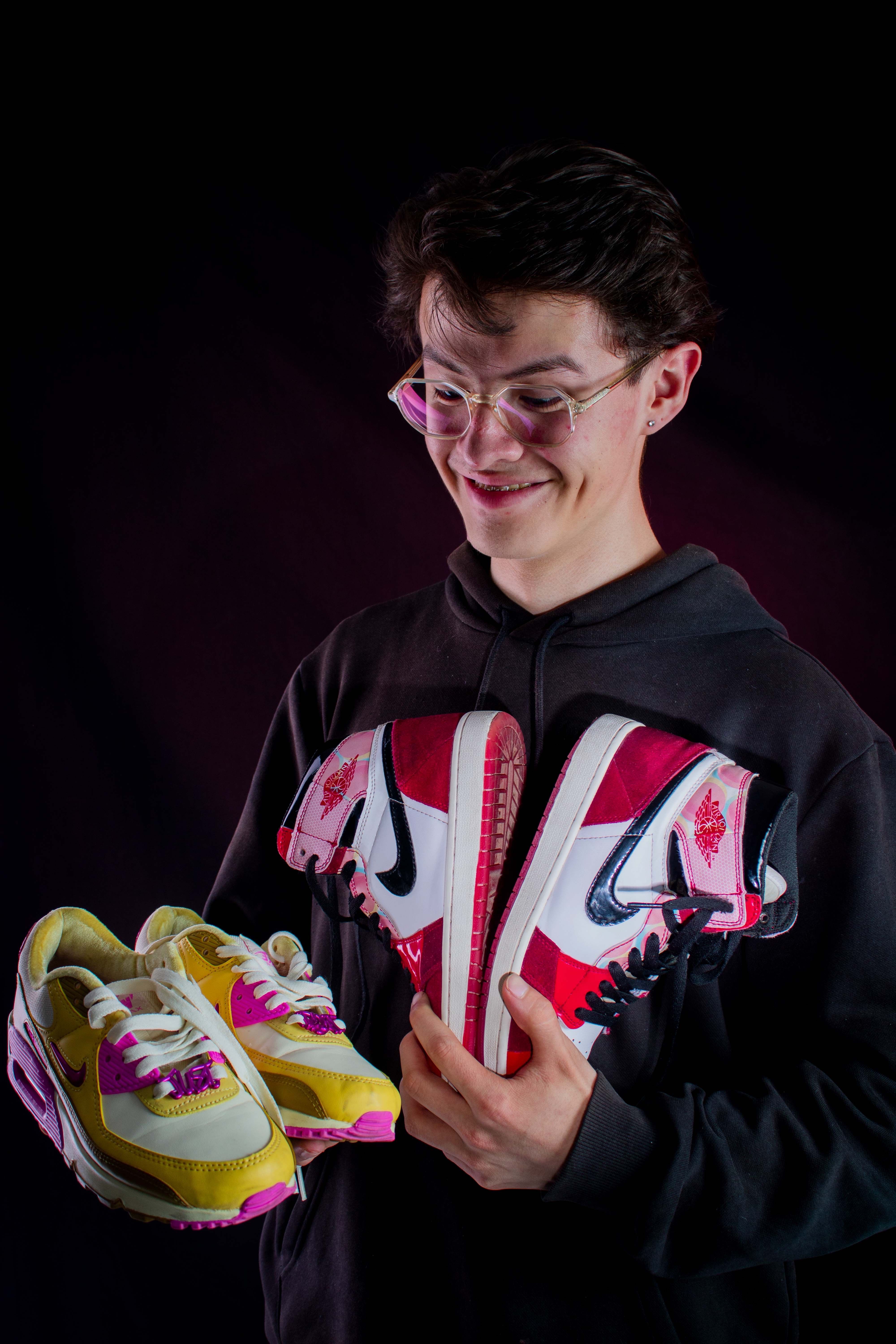 A young man joyfully displays two pairs of vibrant sneakers against a dark backdrop.