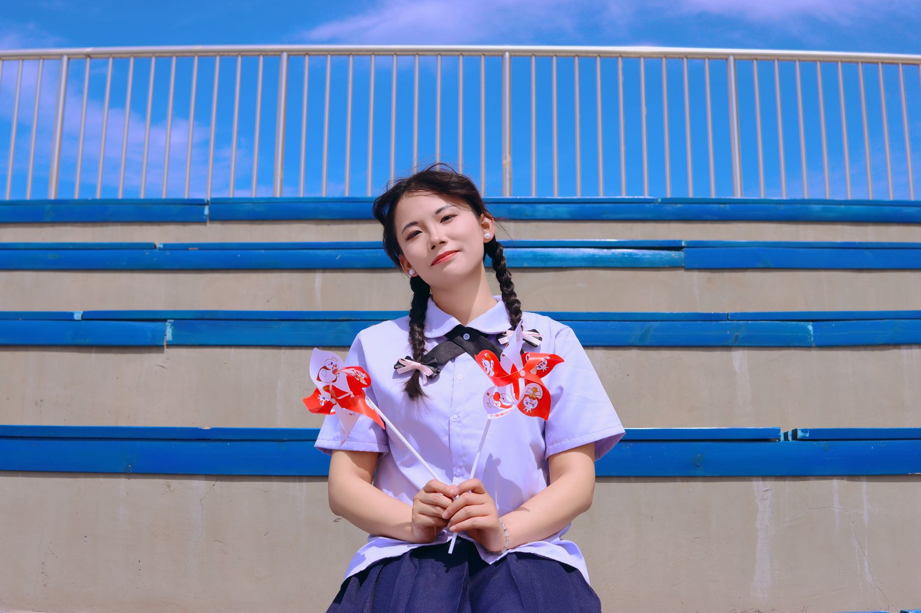 A cheerful young woman holding pinwheels, sitting on outdoor stadium steps.
