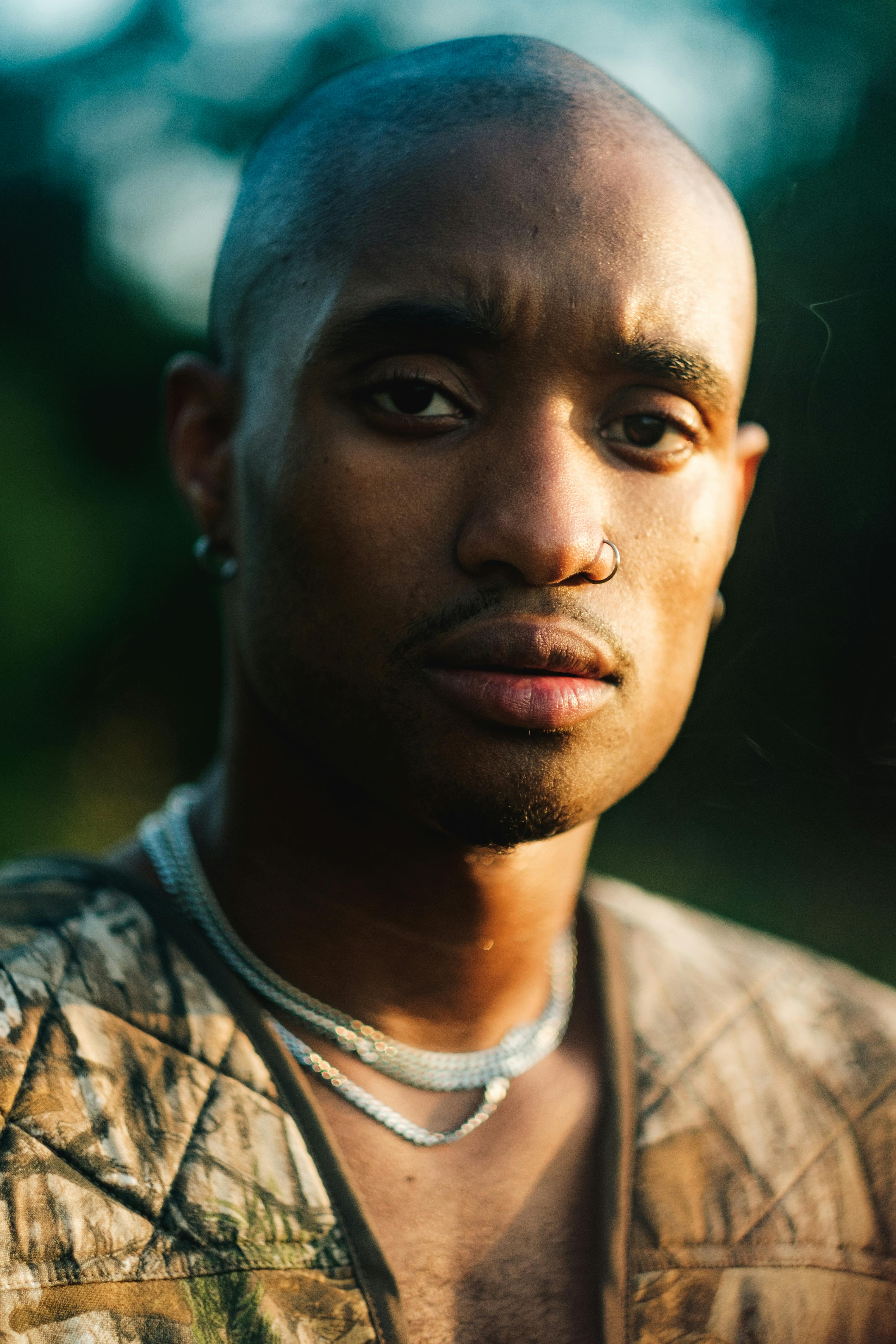 Close-up portrait of a serious bald man with jewelry in soft outdoor lighting.