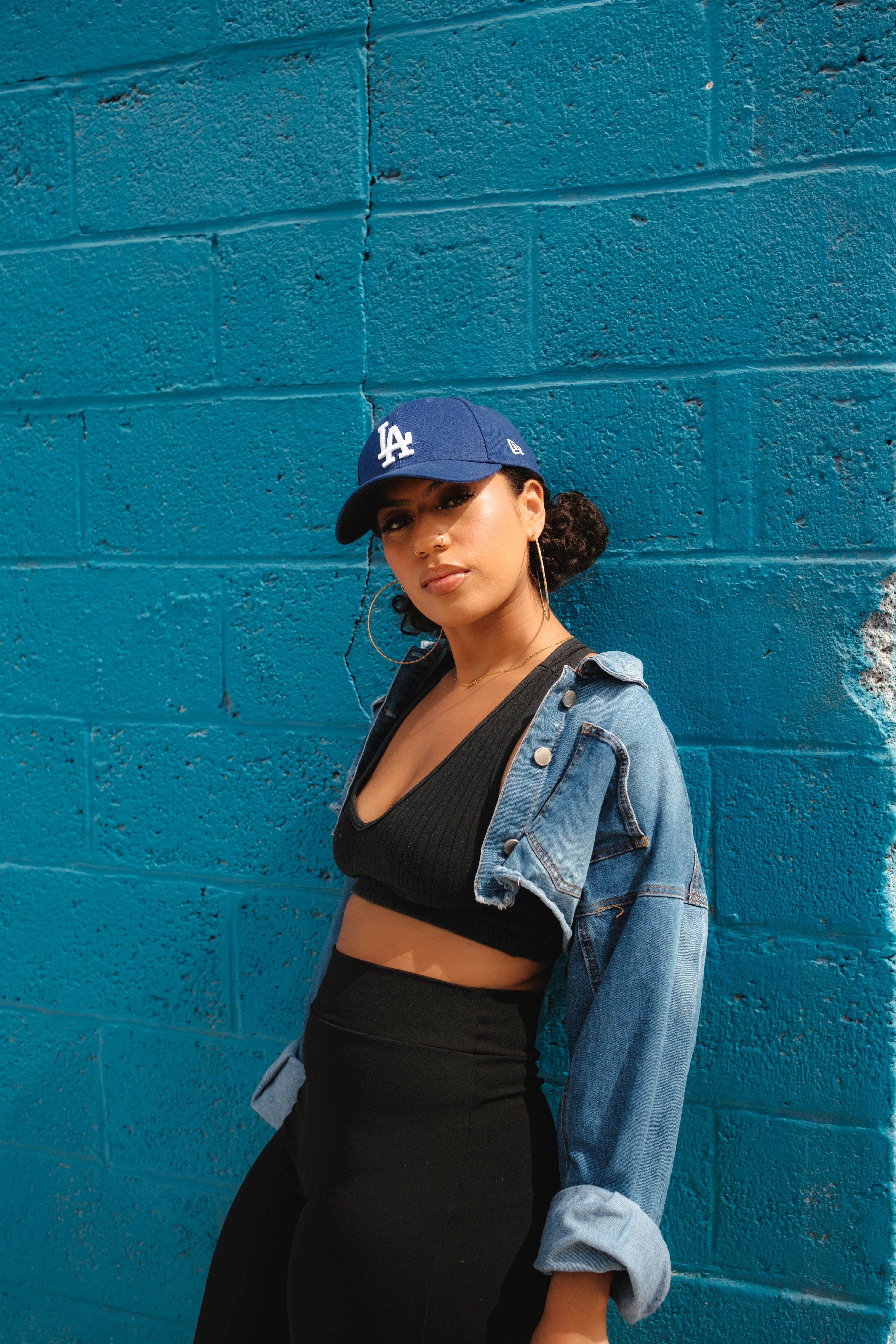 Fashionable young woman in denim jacket and cap posing against a vibrant blue wall, showcasing urban style.