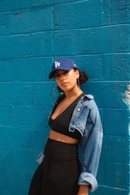 Fashionable young woman in denim jacket and cap posing against a vibrant blue wall, showcasing urban style.