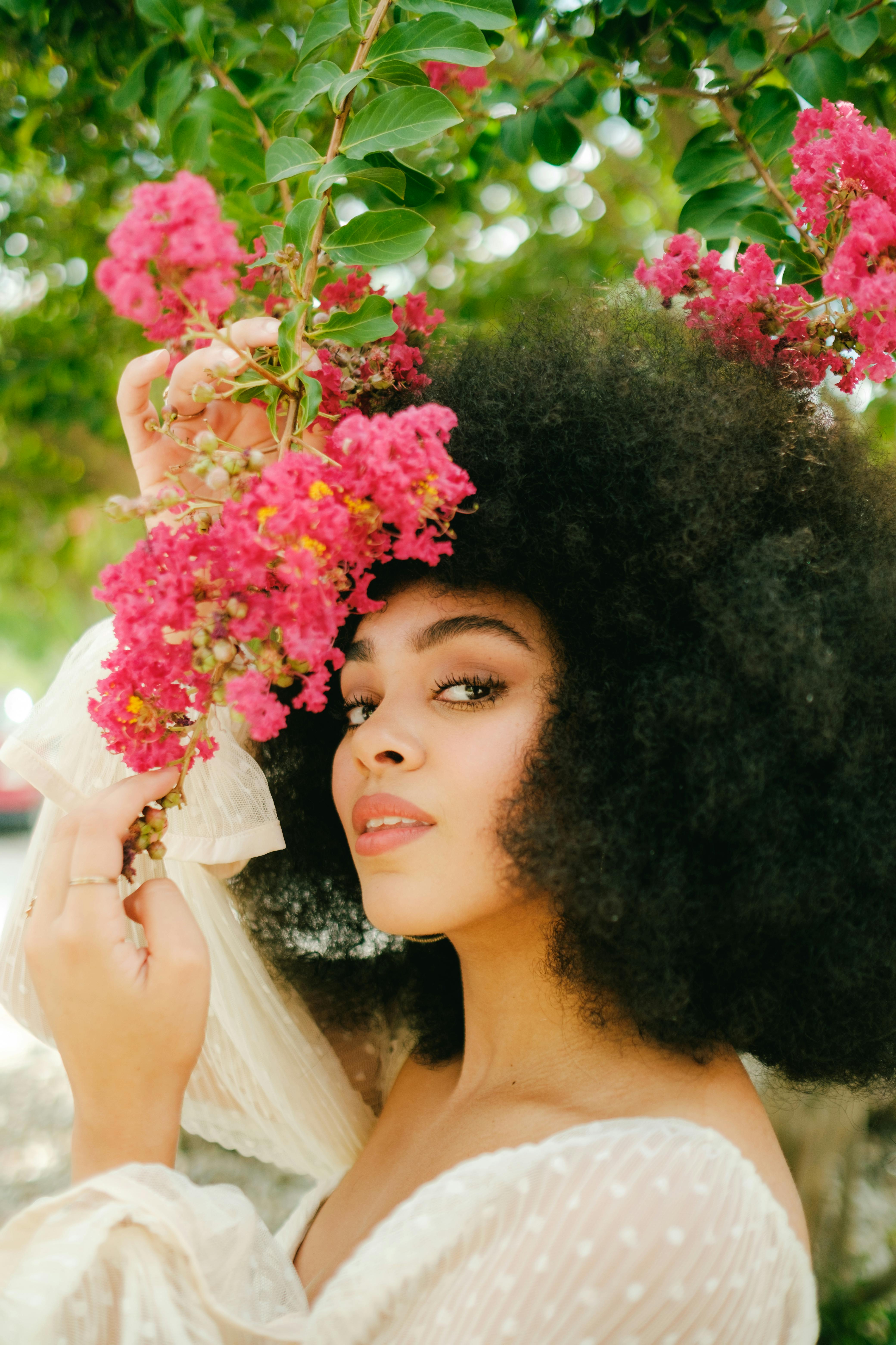 Fashionable woman with afro hairstyle posing among vibrant pink blossoms in a serene outdoor setting.