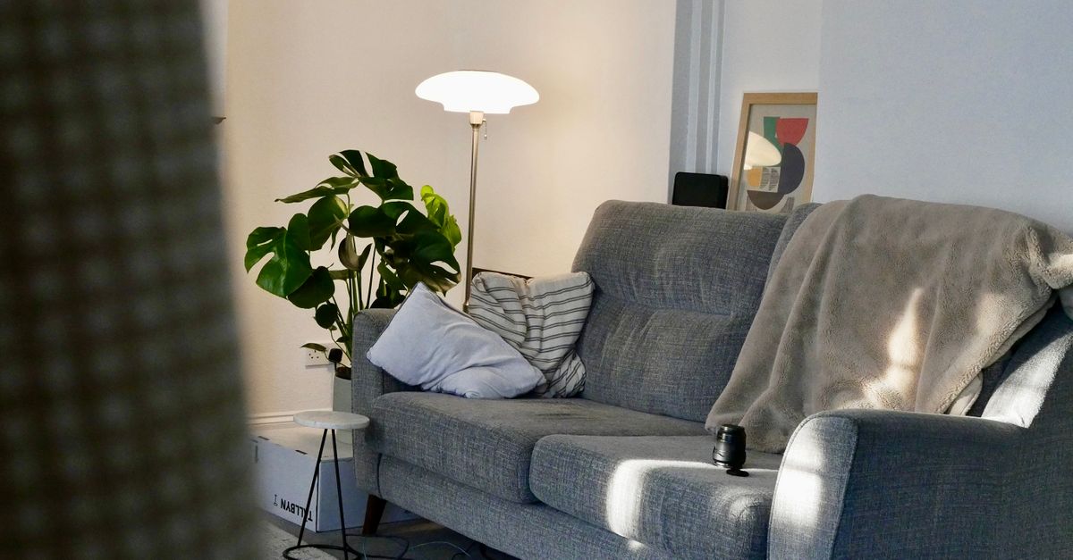 Neben einem grauen Sofa leuchtet eine Stehlampe, auf dem Sofa liegen Kissen und eine Decke