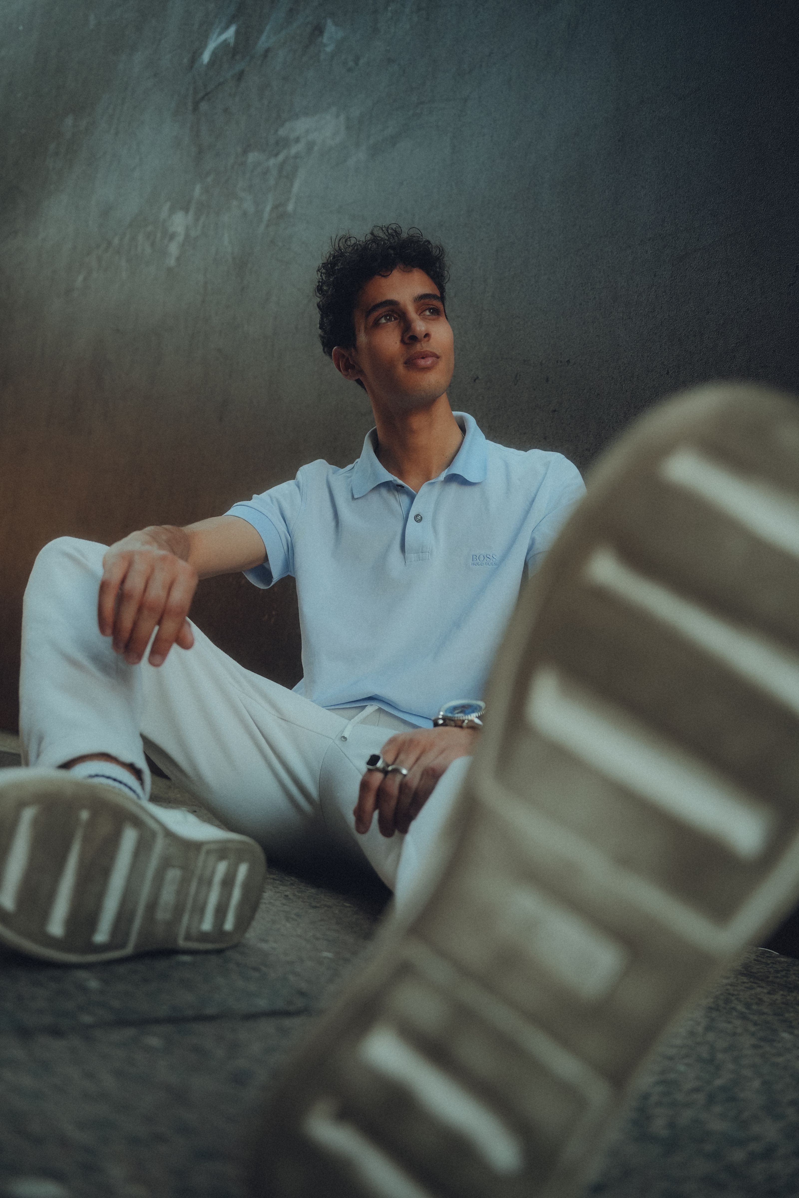 Jovem sentado no chão em um ambiente urbano, vestindo roupas casuais.