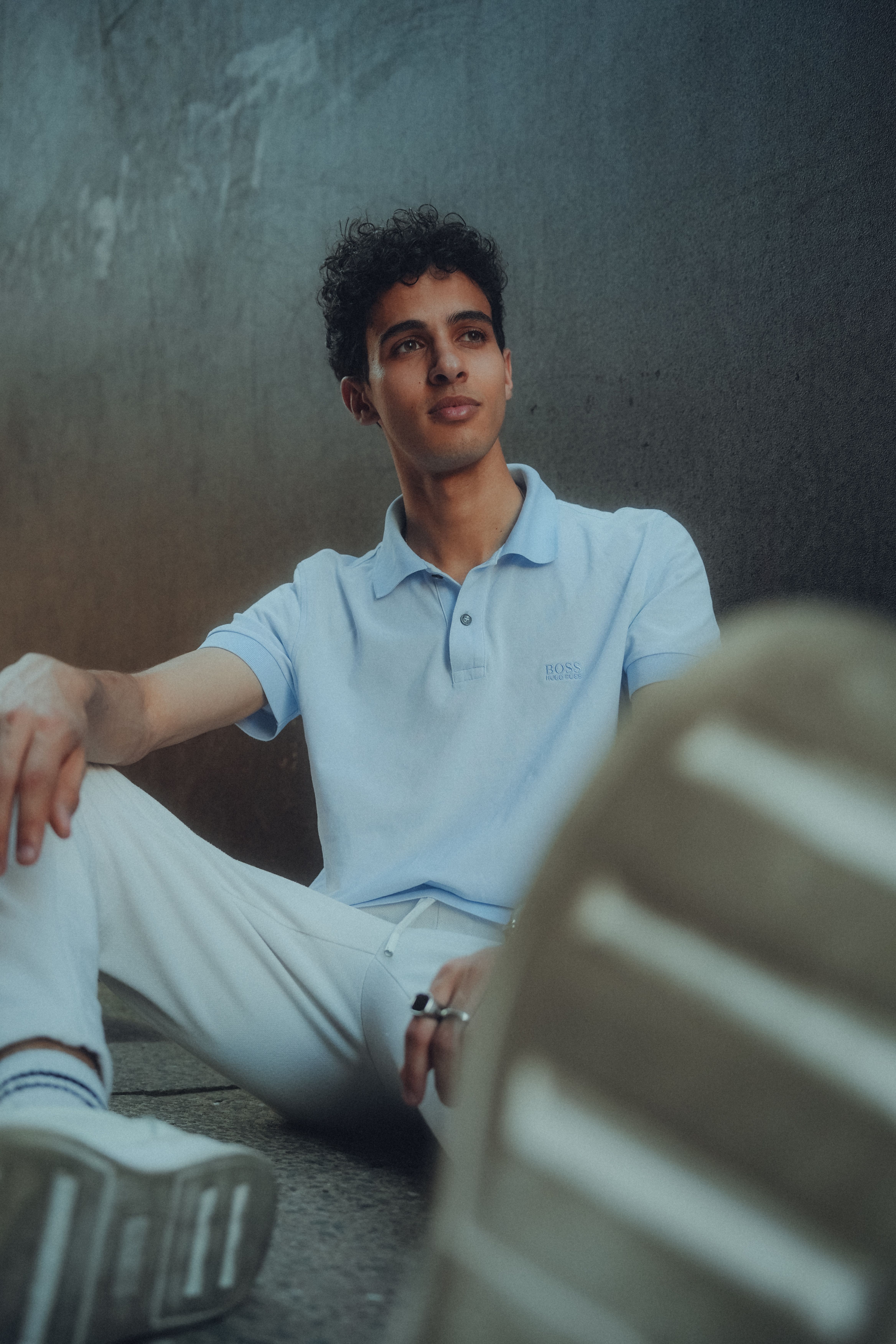 Casual portrait of a young man in a relaxed pose, sitting against a concrete wall in an urban setting.