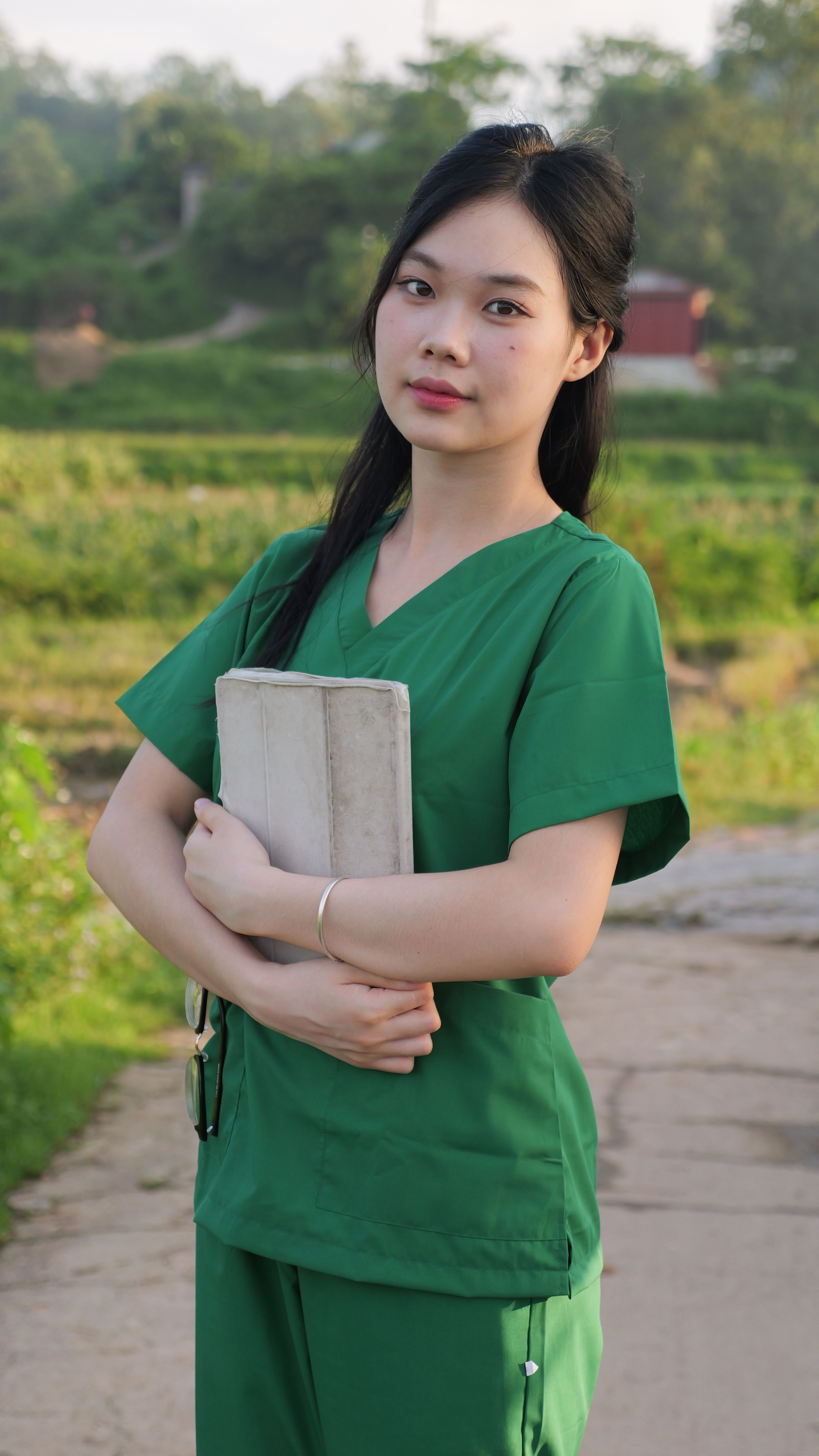 Portrait of a young woman wearing green scrubs holding a book in an outdoor setting.