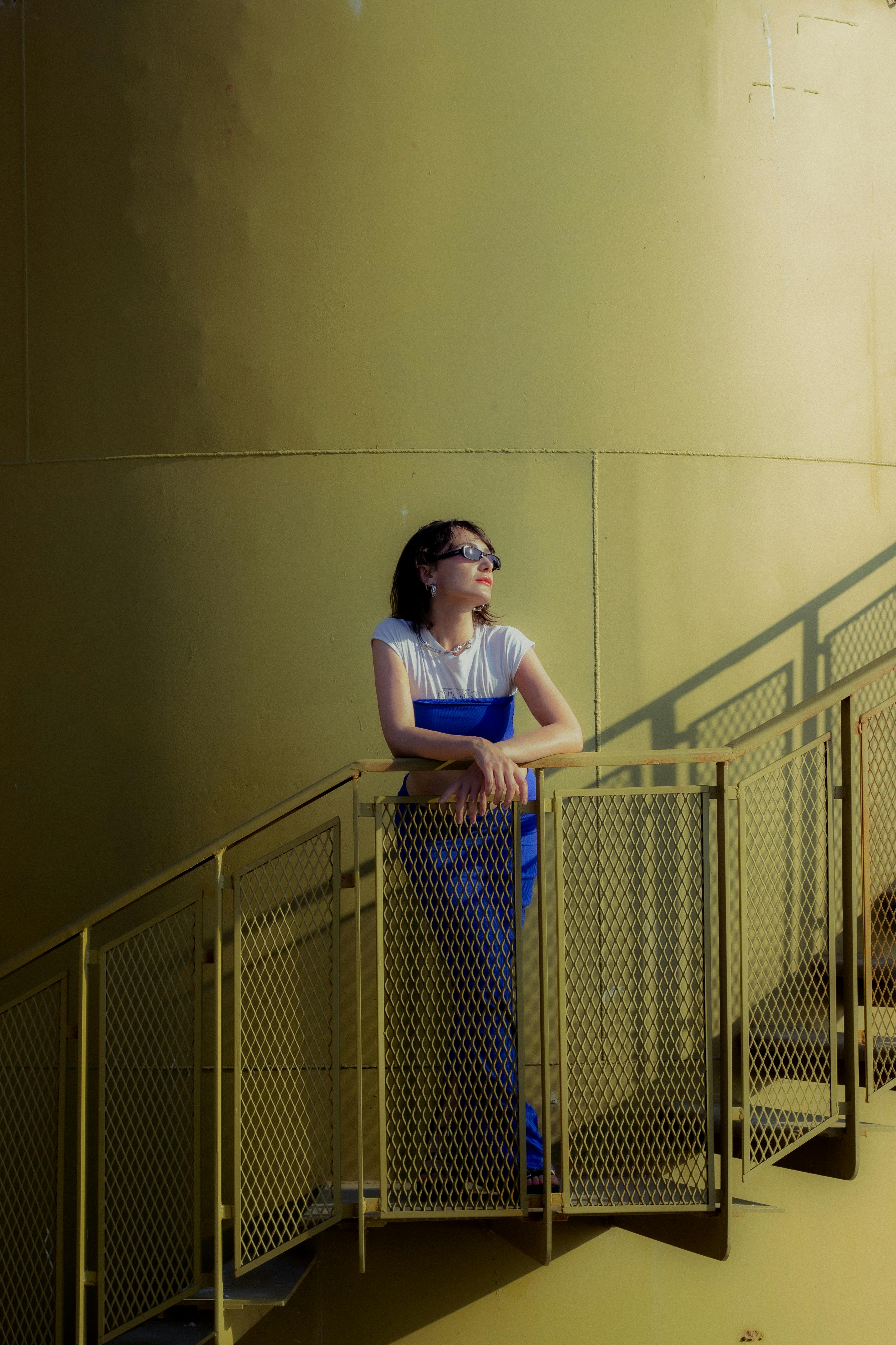 Stylish woman in a blue dress poses on a spiral staircase against a yellow background.