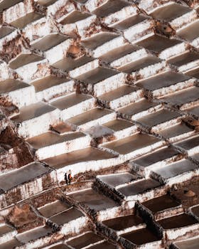 Aerial shot capturing the geometric patterns of salt pans with two people visible, creating a stunning landscape view.
