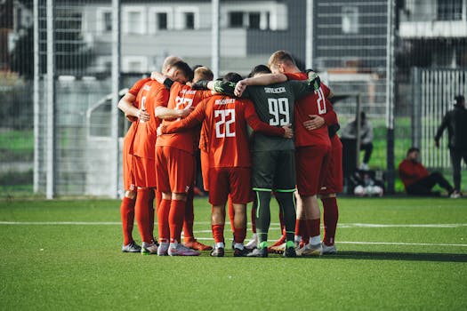 Group of soccer players in red uniforms huddling on a green field before a match.