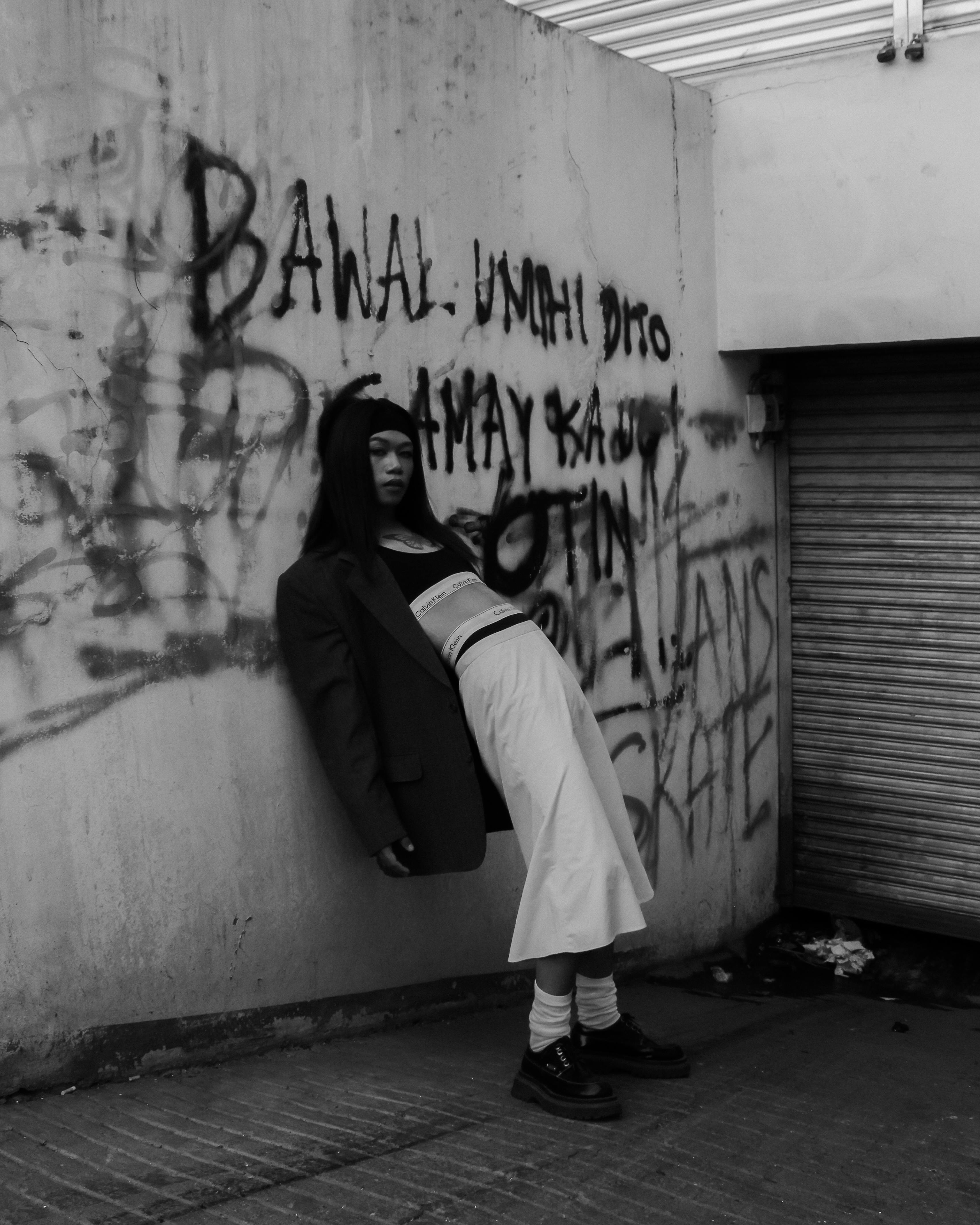 A black and white portrait of a woman leaning against a graffiti-covered wall in an urban setting.