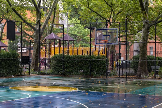 Vibrant urban basketball court next to a playground surrounded by trees on a rainy day.