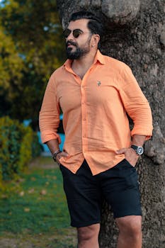 Stylish man in orange shirt and sunglasses relaxing by a tree in autumn park.