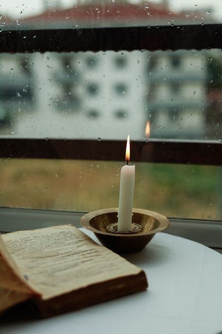 A lit candle and an open book by a rainy window create a serene ambience.