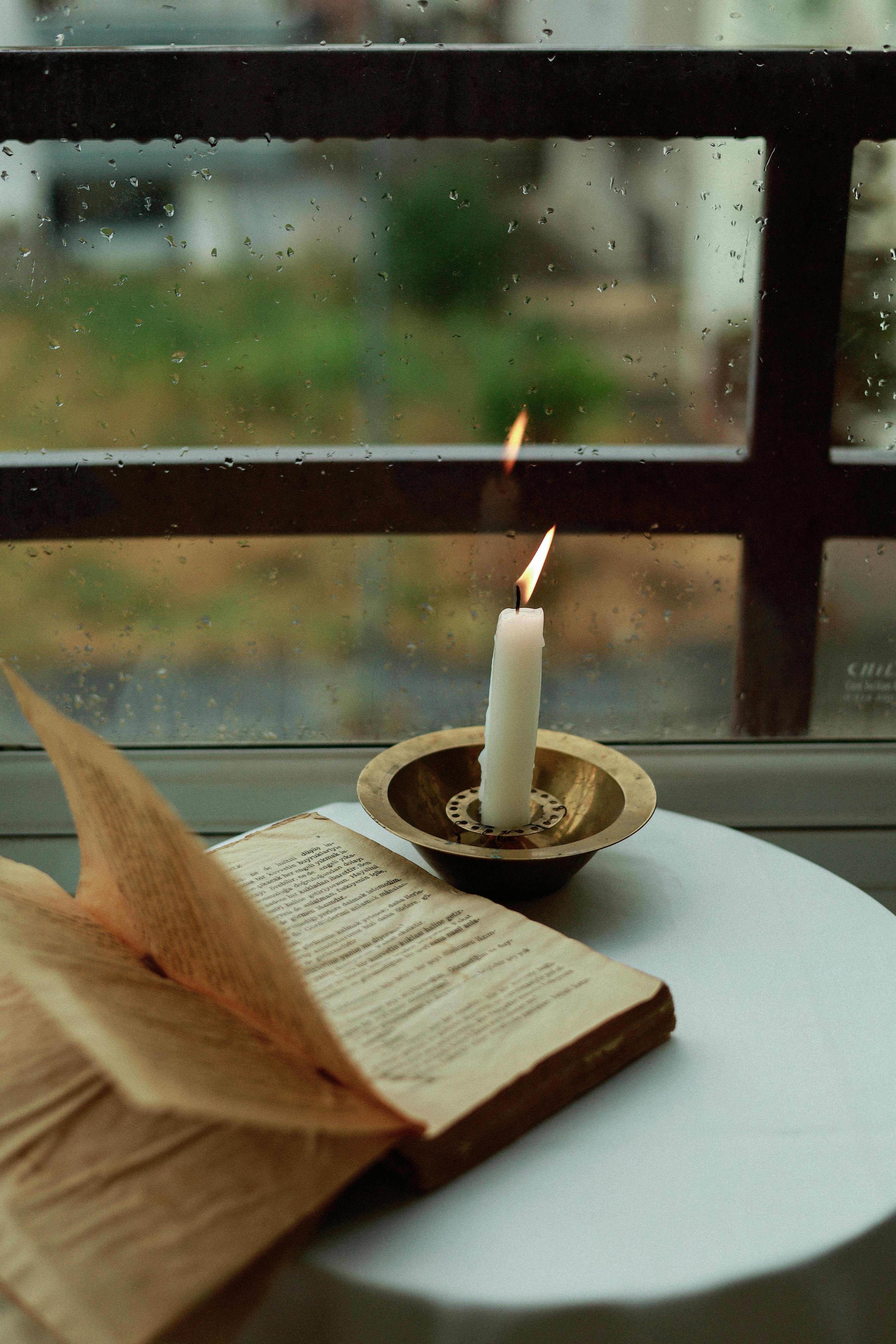 A cozy setup with a candle and open book by a rainy window, perfect for reading.