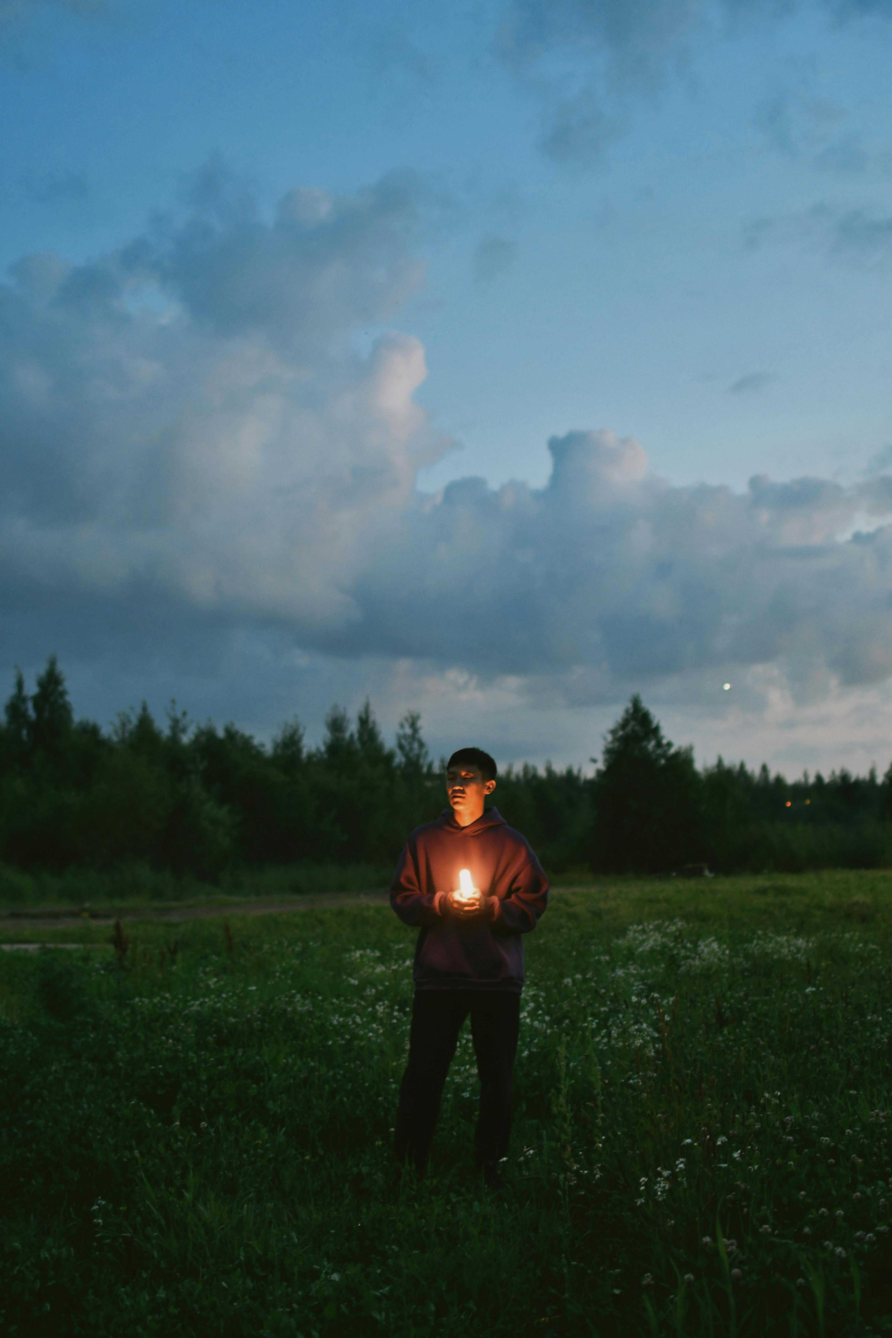 A young man stands in a meadow at twilight holding a glowing candle, surrounded by clouds.