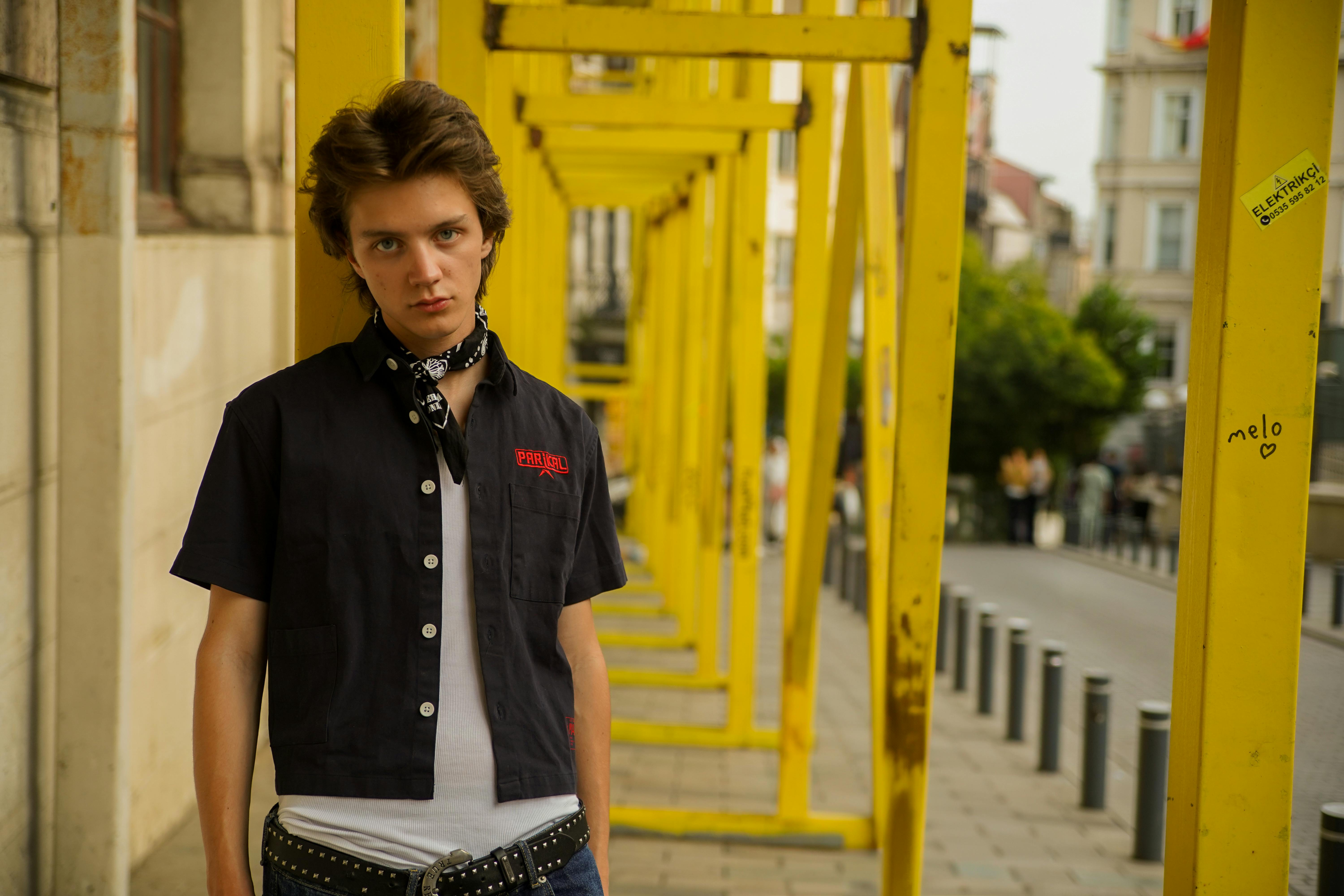 Young male fashion model posing outdoors in an urban setting with yellow construction frames.