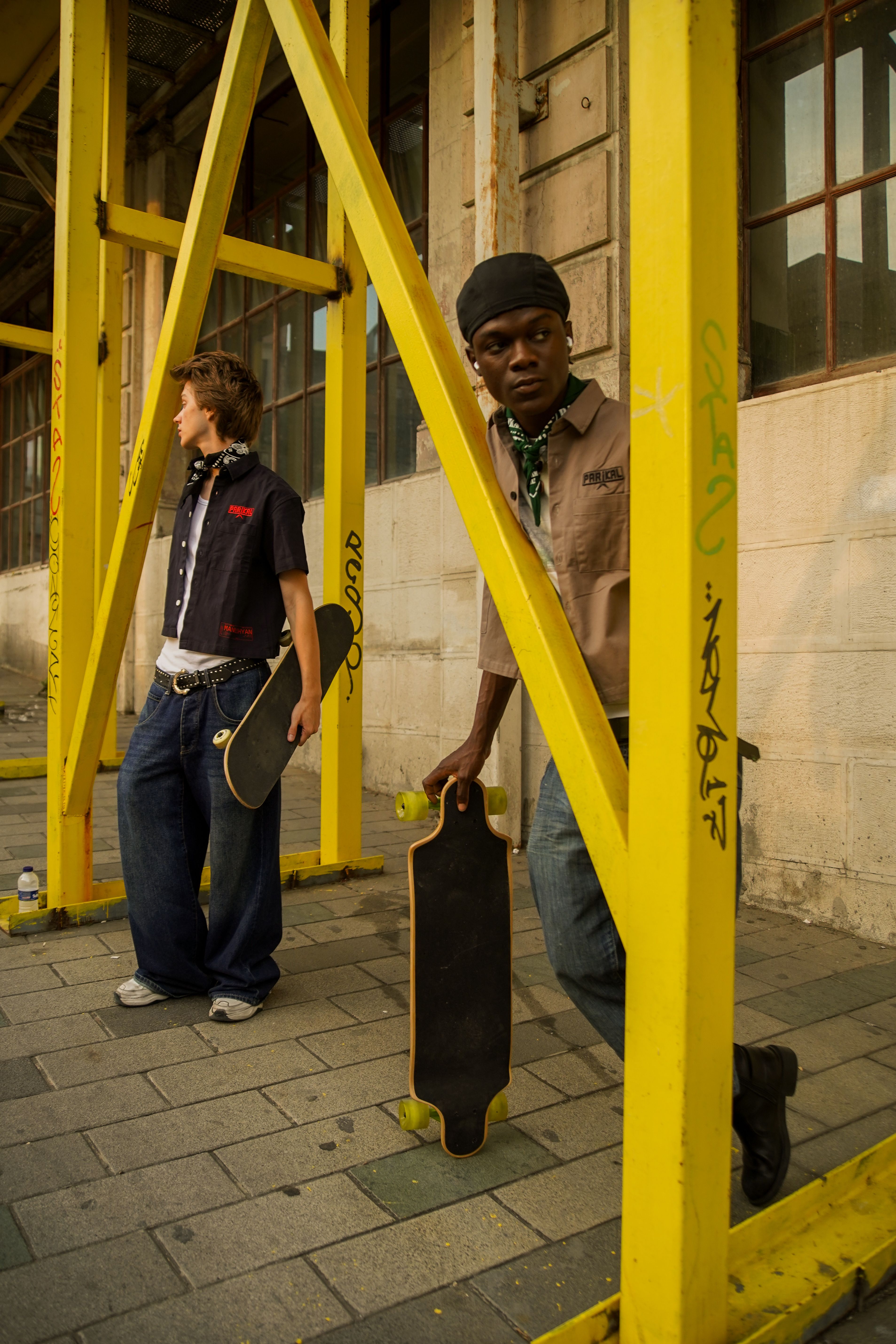 Two skaters with longboards in an urban industrial environment, showcasing street fashion.