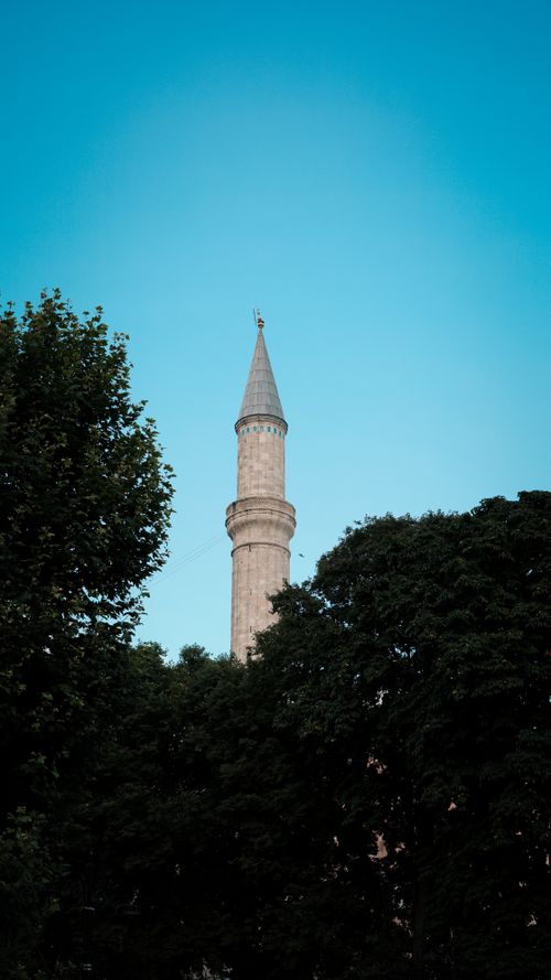 Free A towering minaret rises above green trees against a clear blue sky, symbolizing architecture and spirituality. Stock Photo