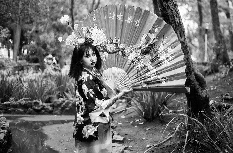 Elegant geisha in kimono with fan standing in a scenic forest park.