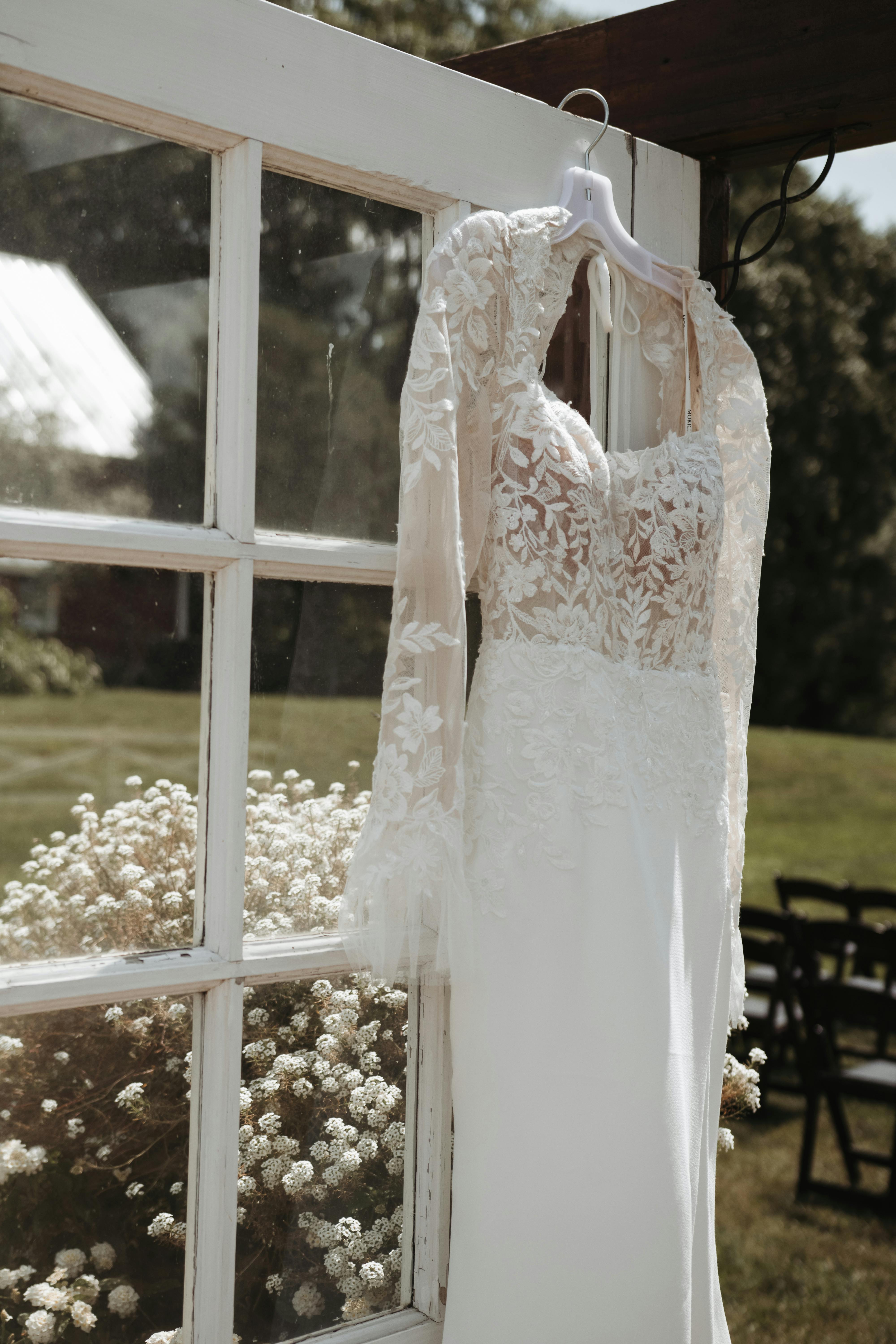 White lace wedding dress hanging by an outdoor window with sunlit background.