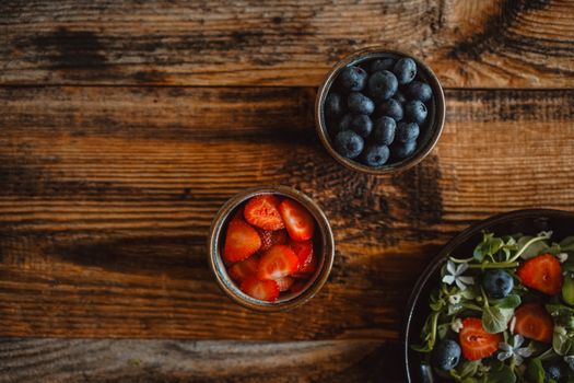 Top view of fresh strawberries, blueberries, and a salad displayed on a rustic wooden table.