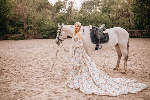 Free A woman in a floral gown leading a white horse on a sandy outdoor arena. Stock Photo