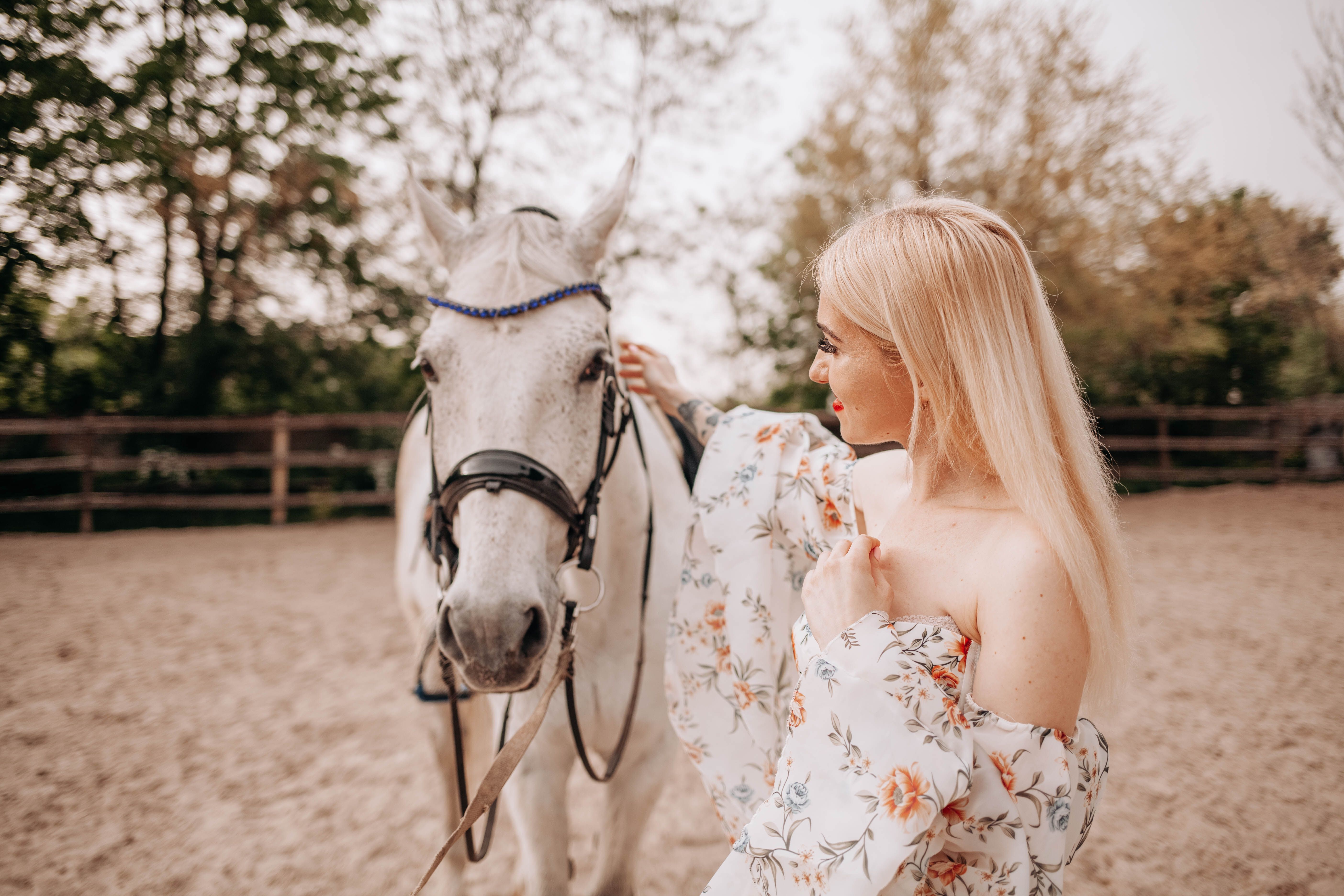 A woman in a floral dress gently interacts with a white horse outdoors in a serene setting.