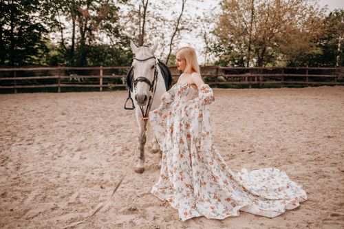 Free A stylish woman in a floral gown standing next to a white horse in a sandy outdoor setting. Stock Photo