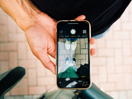 A person captures shoe reflection on a smartphone, showcasing modern technology and leisure outdoors.