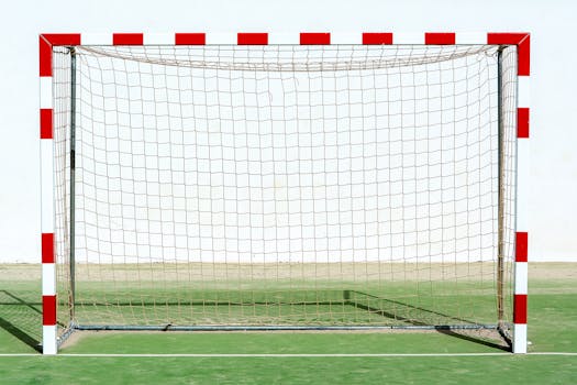 A solitary soccer goal with red and white colors on an outdoor sports field against a plain background.