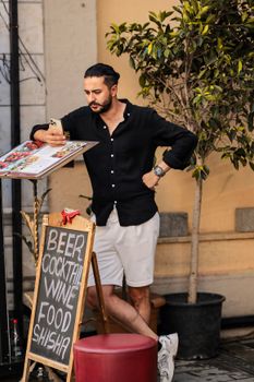 Young man in casual wear checking a menu at an outdoor cafe with drinks menu display.