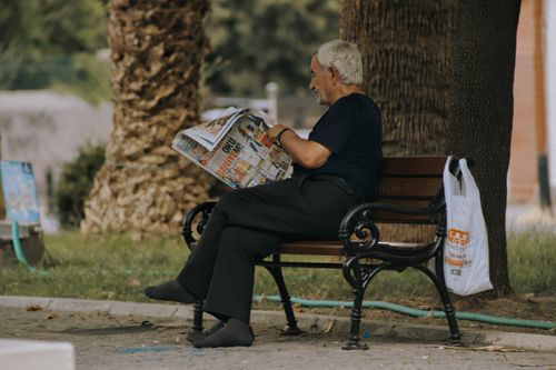 Free A senior man in a park enjoys reading a newspaper while sitting on a bench. Stock Photo