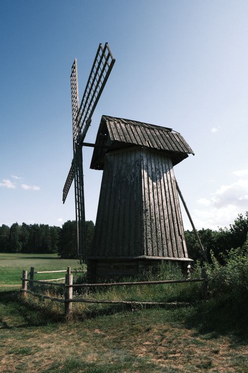Free A rustic wooden windmill stands amidst a rural landscape under a clear blue sky. Stock Photo