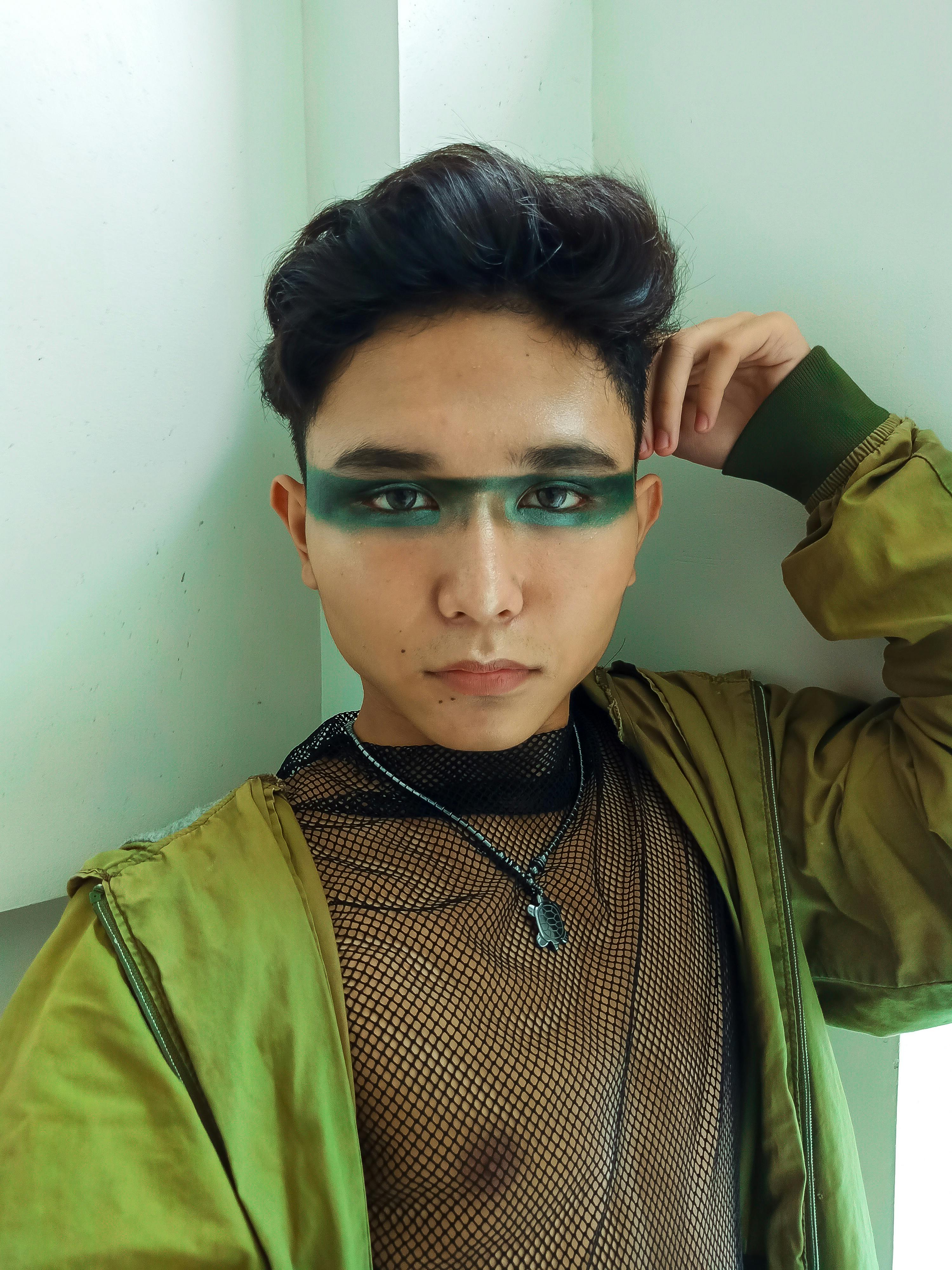 Portrait of a fashionable individual with painted eyes and a modern green jacket indoors.