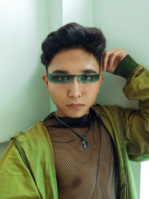 Portrait of a fashionable individual with painted eyes and a modern green jacket indoors.
