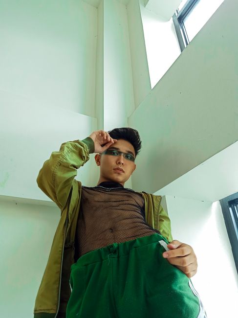 Low angle shot of a fashionable young man indoors with striking eye makeup and casual attire.