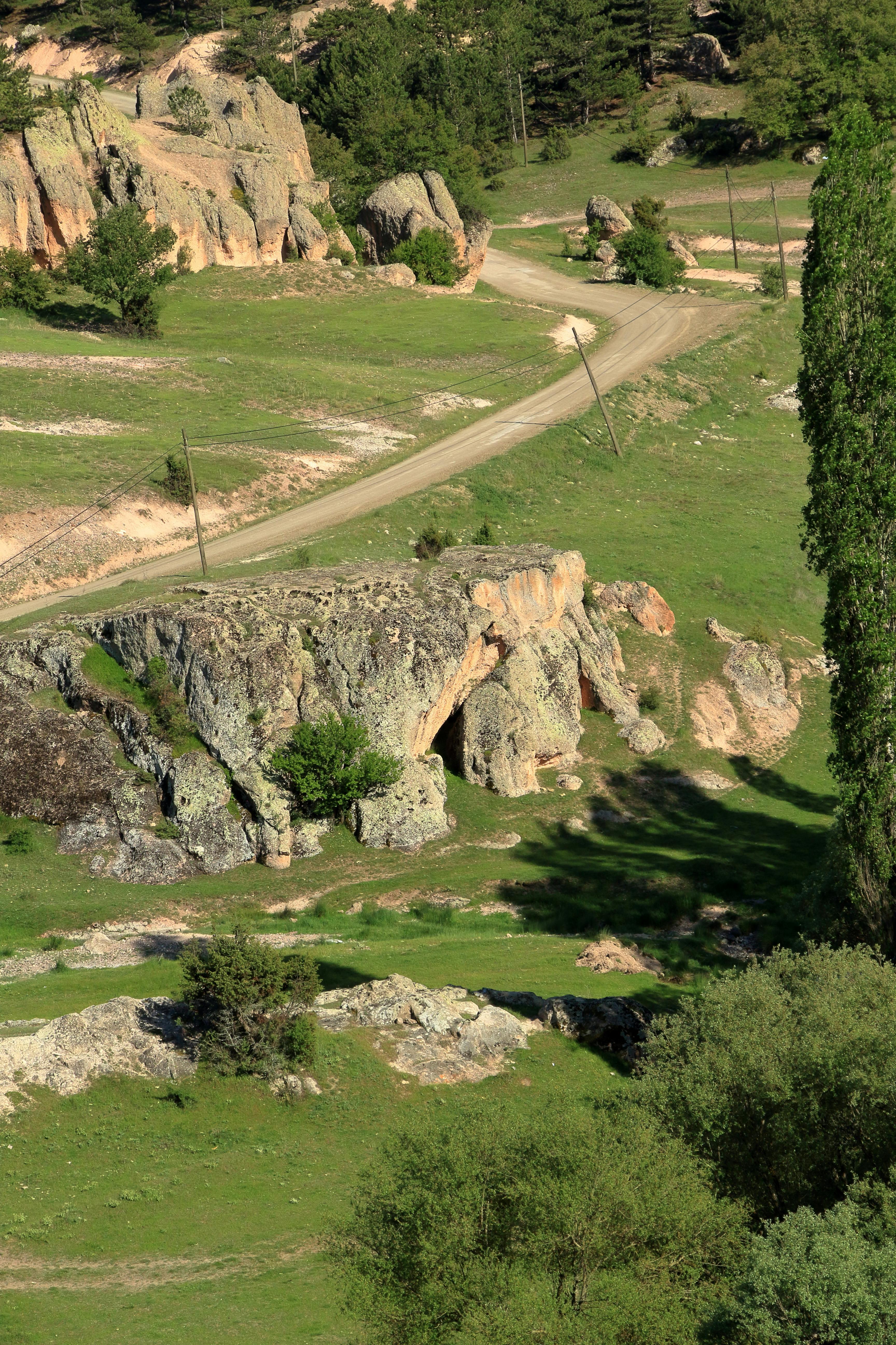 A winding road meanders through a lush, rocky countryside landscape on a sunny day.
