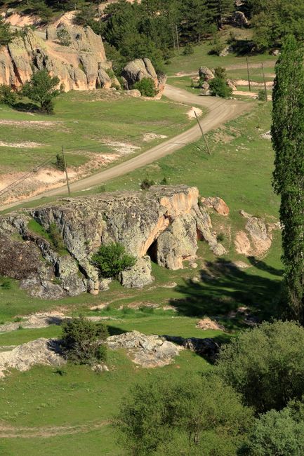 A winding road meanders through a lush, rocky countryside landscape on a sunny day.