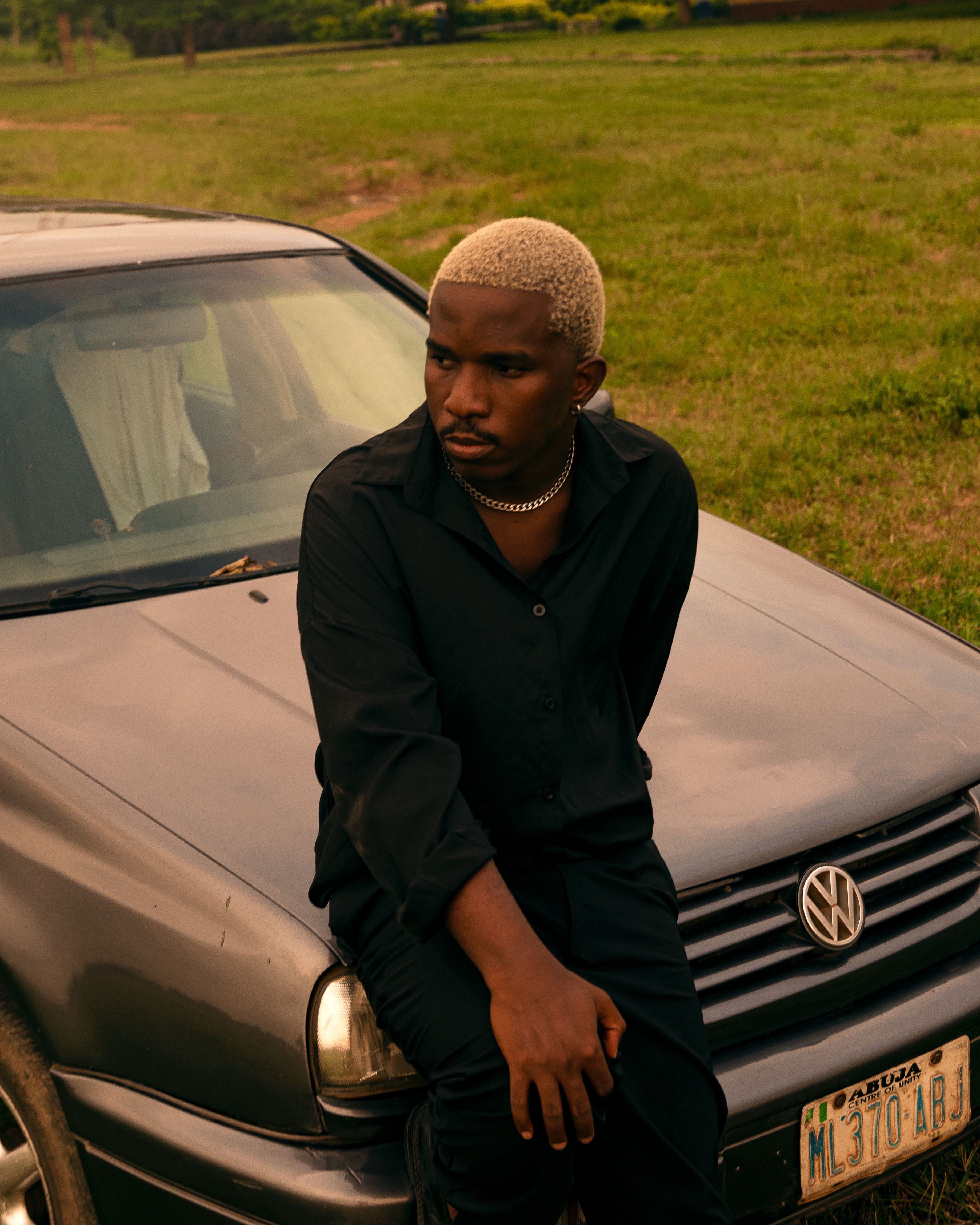 A man with blonde hair sits on a car hood in a grassy area under warm light.
