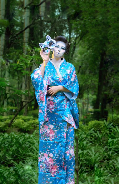 Free A woman in a blue kimono holding a mask, standing in a lush forest setting. Stock Photo