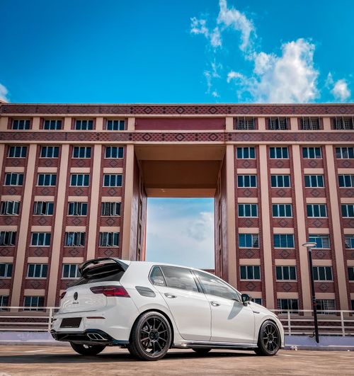 Free White Volkswagen Golf parked in front of a contemporary urban building under a blue sky. Stock Photo