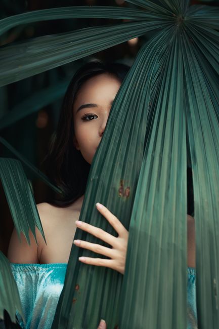 A woman elegantly poses behind tropical leaves, creating a mysterious and stylish feel outdoors.