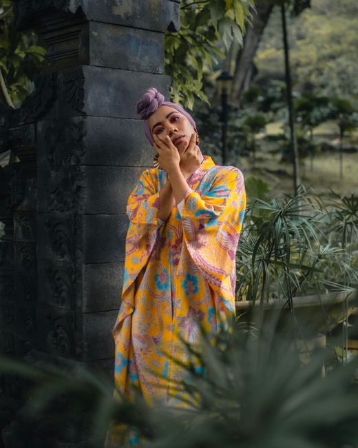 Fashionable woman posing in vibrant dress and headscarf amidst lush greenery.
