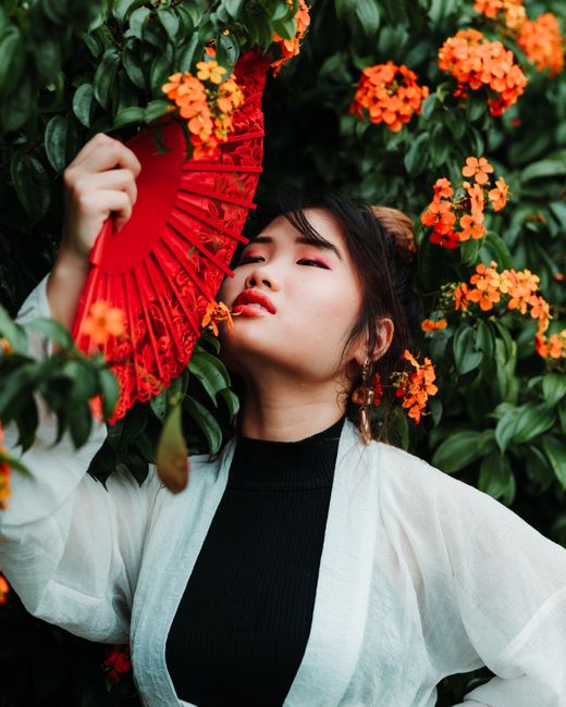 Artistic outdoor portrait of a woman with a red fan amidst vibrant orange flowers in Kuala Lumpur.