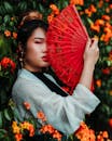 一位时尚的亚洲女性手持传统红扇，在鲜艳的花丛中摆出姿势